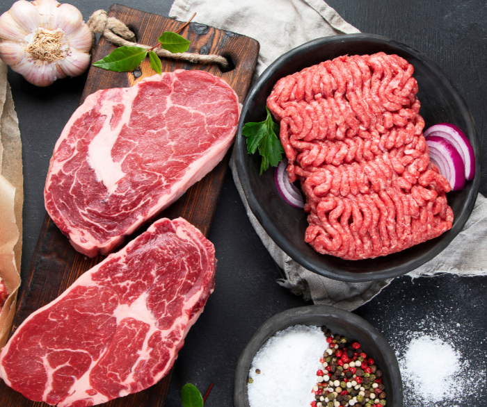 Fresh cuts of beef including ribeye steaks and ground beef, garnished with garlic, herbs, and spices on a dark rustic background.
