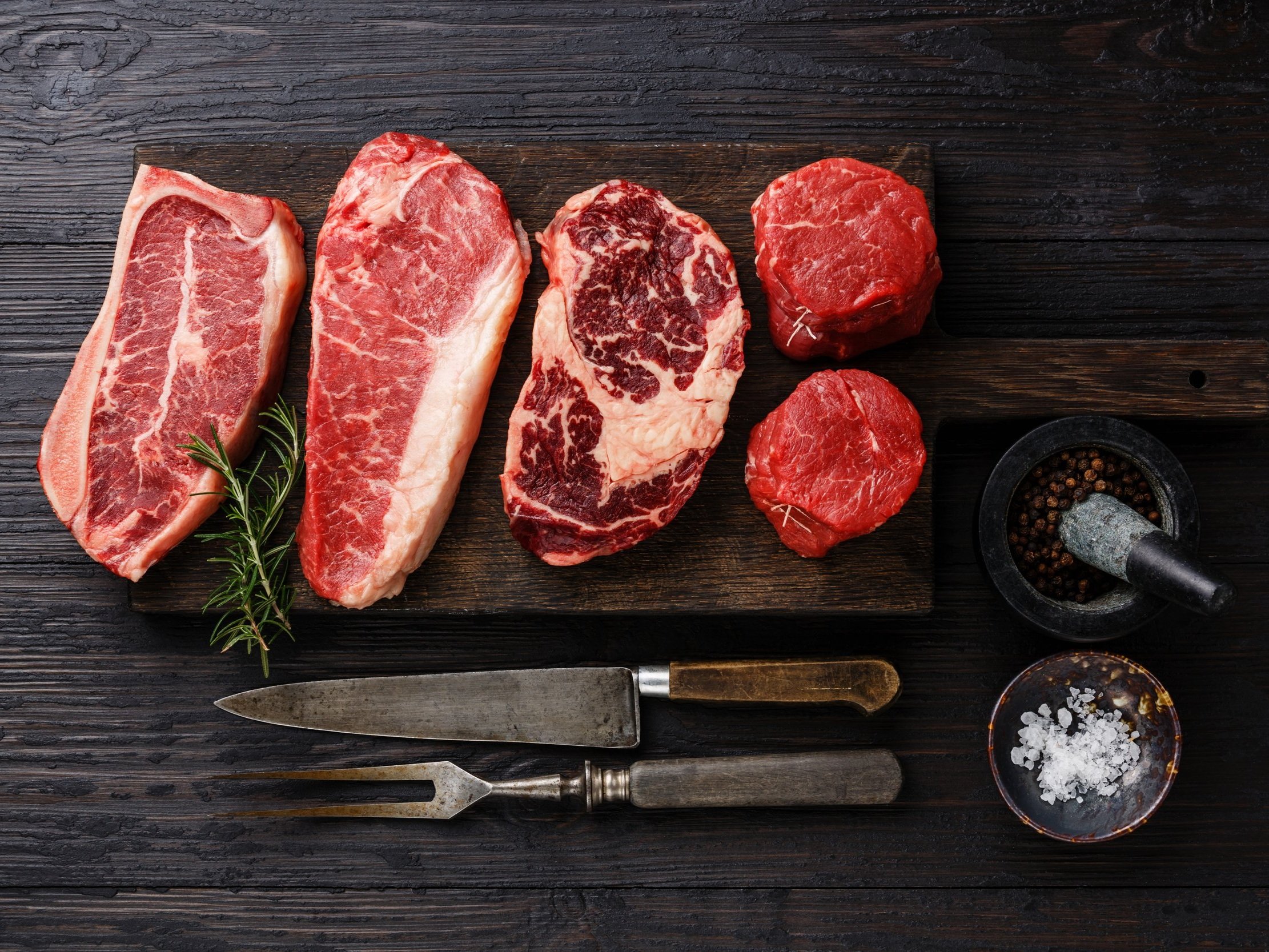 Assorted cuts of raw beef including ribeye, sirloin, and tenderloin on a wooden cutting board, accompanied by fresh rosemary, a mortar and pestle with peppercorns, and a bowl of sea salt, perfect for grilling or cooking.